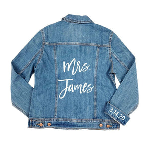 A jean jacket lays showing the customized last name on the back and the custom date adhered to the cuff.