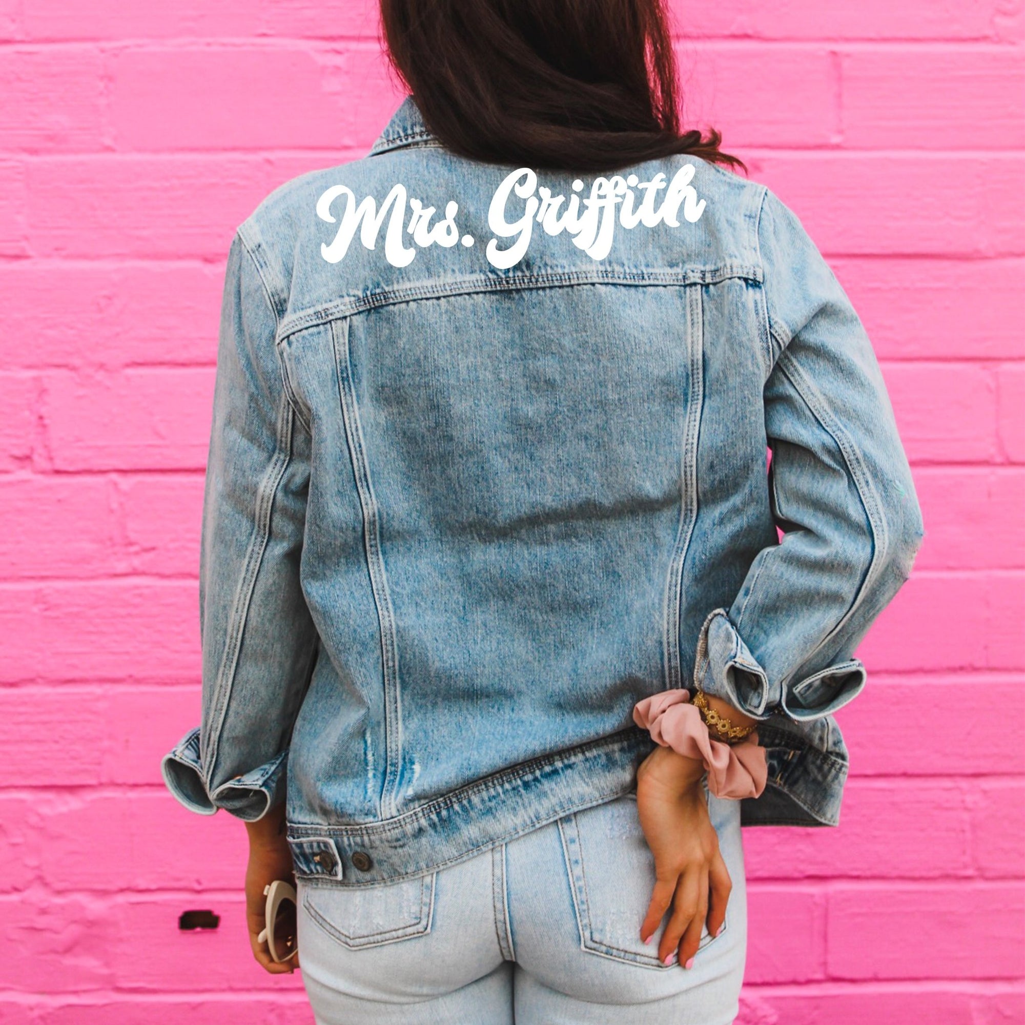 A woman in front of a pink wall wears a jacket which reads "Mrs. Griffith" across the shoulders.