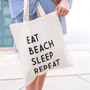 A woman holds a tote bag which reads "Eat Sleep Beach Repeat."