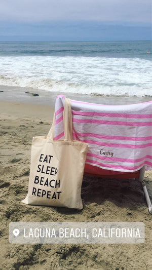 A beach chair is set up in front of the water with a customized towel and a "Eat Sleep Beach Repeat" tote draped over it.