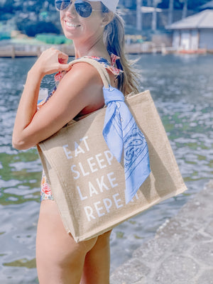 A woman stands by a lake holding her "Eat Sleep Lake Repeat" Jute Tote with a tied blue bandana over her shoulder.
