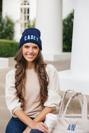 A brunette holds a coffee while wearing an embroidered beanie