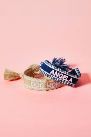A blue bracelet and a beige bracelet are stacked in front of a  pink background showing off their embroidered customizations which read "Dog Mom" and "Angela."