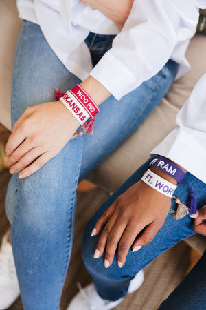 Two women sit with their hands resting on their legs with their custom embroidered bracelets that show off their school pride.