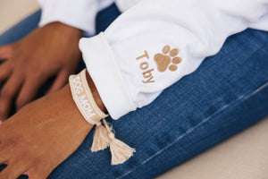 A woman wears a white sweatshirt with an embroidered sleeve which has a beige paw print and the name "Toby"  and a beige bracelet which reads "Dog Mom."