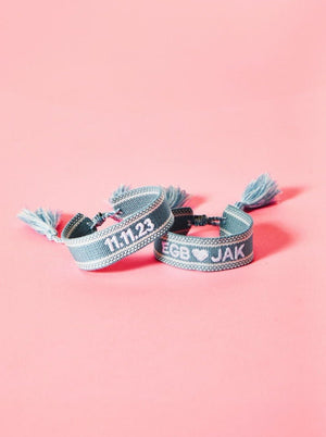 Two light blue bracelets are customized with an embroidered date and two monograms with a heart in between them.