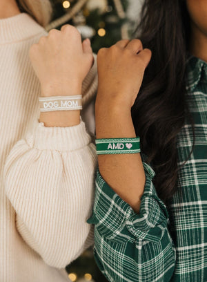 Two women hold up their wrists to show off their green and beige custom embroidered bracelets.