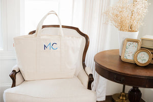 A natural colored canvas tote is embroidered with a blue monongram and placed onto a white chair.