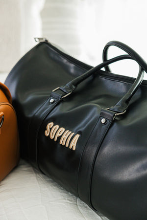 A black duffel bag is personalized with a name in a tan and beige thread.