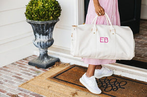 A person walks into an entryway holding a white duffel with a pink monogram.