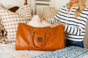 A woman opens a tan duffel with a navy monogram on a bed.