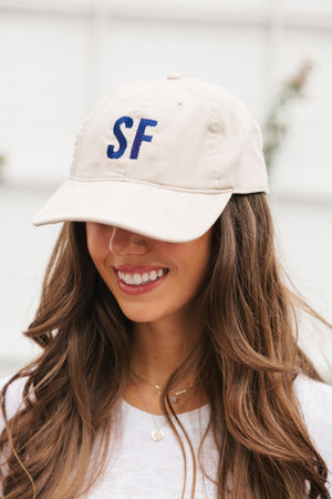 A smiling brunette wears a cream colored monogramed baseball hat