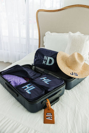 A suitcase is laid open on a bed with a set of navy packing cubes, a navy nylon pouch, and a straw hat which are all embroidered with a monogram.