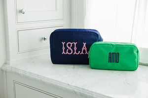 A navy blue nylon pouch is embroidered with a pink name and a green nylon pouch is embroidered with a navy monogram.