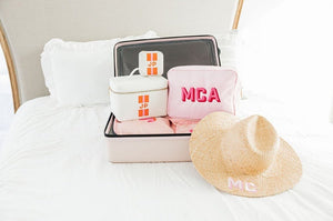 A suitcase is laid on a bed showing a nylon pouch, a train case, a jewelry case, and a straw hat which have all been monogrammed.