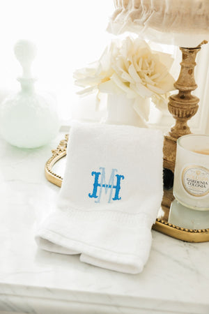 A white hand towel is placed on a bathroom tray and is personalized with a blue monogram.
