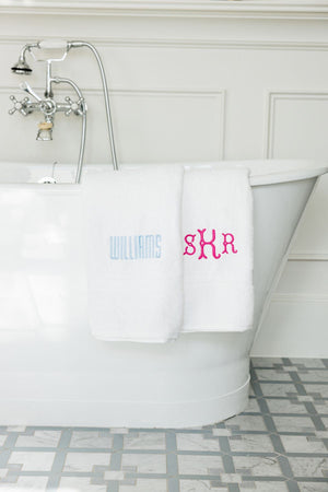 Two white towels lay draped over a bathtub with a blue last name and a pink monogram embroidered on them.