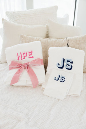 A towel with a pink monogram is wrapped in a pink velvet bow and placed next to a set of bath and hand towels that are monogrammed with blue thread colors.