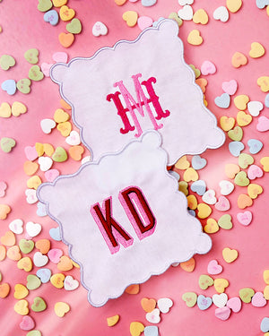 Two scalloped cocktail napkins are monogrammed with pink and red embroidery
