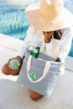A girl in a straw hat holds a navy cooler tote with a green monogram and a green wine glass at the pool.