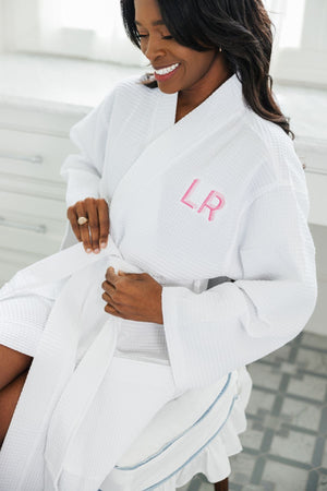 A woman ties a knot in her white waffle knit robe with a pink monogram on it.