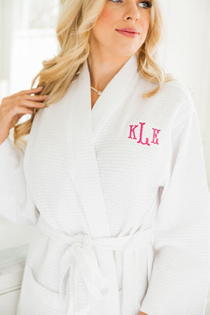 A blonde woman wears a white waffle knit robe with a pink monogram on it.