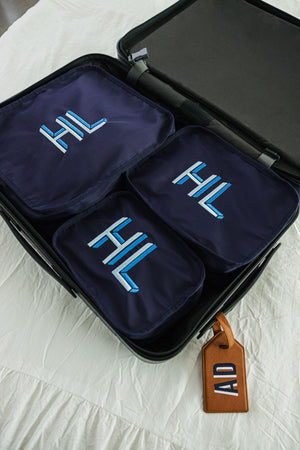 A set of three navy packing cubes with light blue monograms lay in a suitcase.