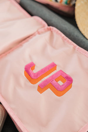 A pink and orange monogram is embroidered onto a pink packing cube.