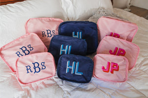 A group of pink and navy packing cubes are embroidered with monograms and placed on a bed
