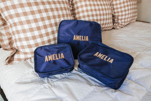 A group of navy packing cubes are embroidered with a name in beige and tan threads.