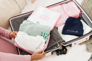 A person packs up their custom embroidered sweatshirts in a pink seersucker packing cube next to a pink suitcase and a navy beanie which reads "montauk."