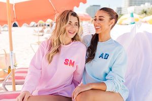 2 women sit on the beach wearing custom sweatshirts in pink and blue