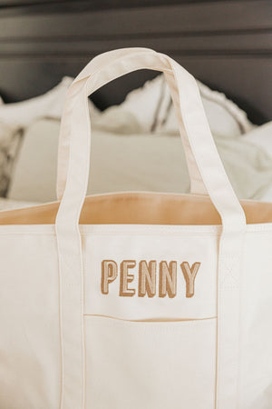 A cream tote bag is embroidered with a name in beige and tan thread.