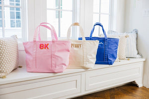 A pink, cream, and a navy tote bag are personalized with names and monograms and placed on a bench.