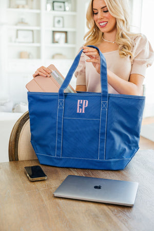 A woman places her electronics into her blue tote bag with a pink embroidered monogram.