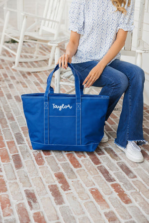 A woman sits on a patio with a blue tote bag which is personalized with a name in a light blue thread color.