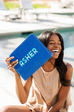 A woman sits and laughs holding up a blue pouch with "Asher" embroidered on it.