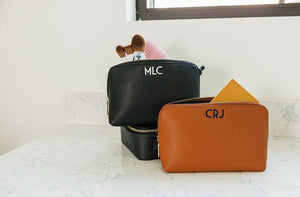 A tan and a black leather pouch are embroidered with monograms and filled with beauty products.