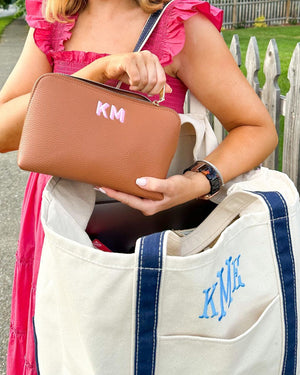 A woman holds a tan leather pouch with a pink monogram and a blue canvas tote with a blue monogram.