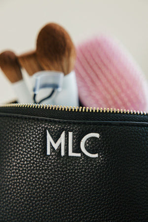 A black leather pouch is embroidered with a white monogram and filled with makeup brushes.