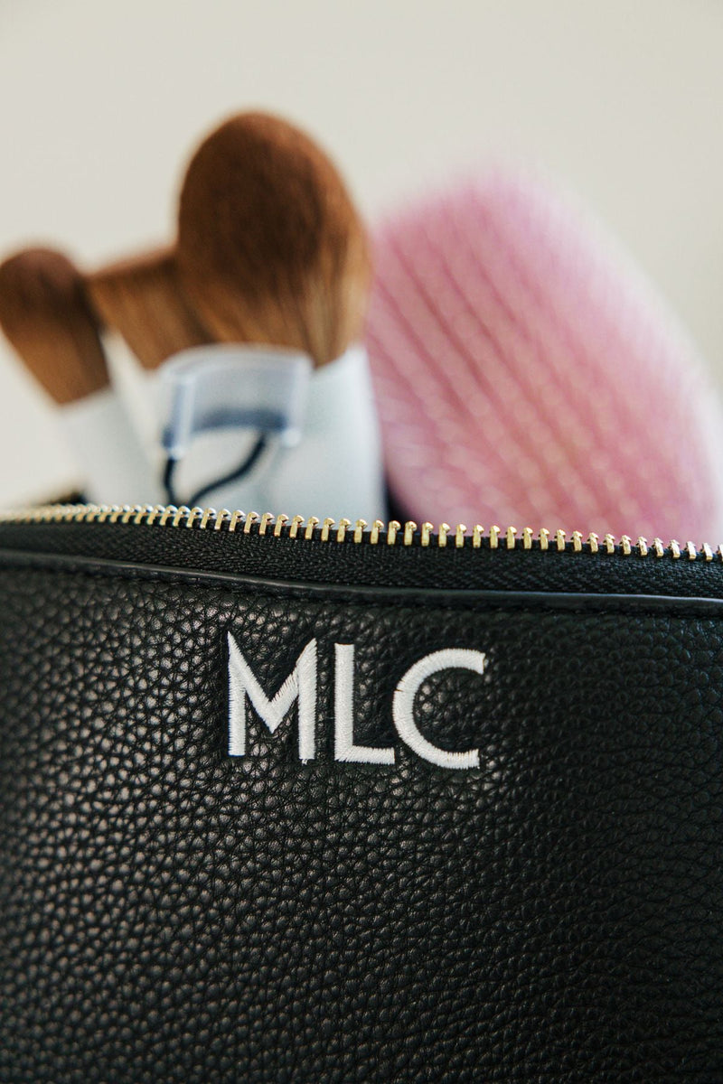 A black leather pouch is embroidered with a white monogram and filled with makeup brushes.