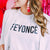 A white t shirt lays flat and reads "Feyonce" in black, block font.