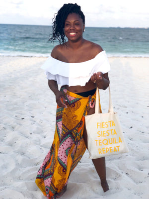 A woman stands on the beach holding her tote which says "Fiesta Siesta Tequila Repeat" in yellow.