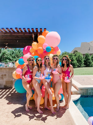 A group of women stand at the pool in front of some balloons posing with their flamingo drink floats.