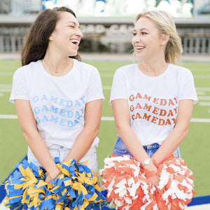 Two girls smile and laugh with each other wearing customized gameday shirts and holding matching pom poms