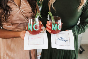 Two women hold cocktail napkins under their drinks which are embroidered with custom text.