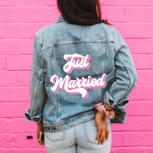 A woman stands in front in a pink wall showing off her customized denim jacket with "Just Married" printed on it in white and pink.