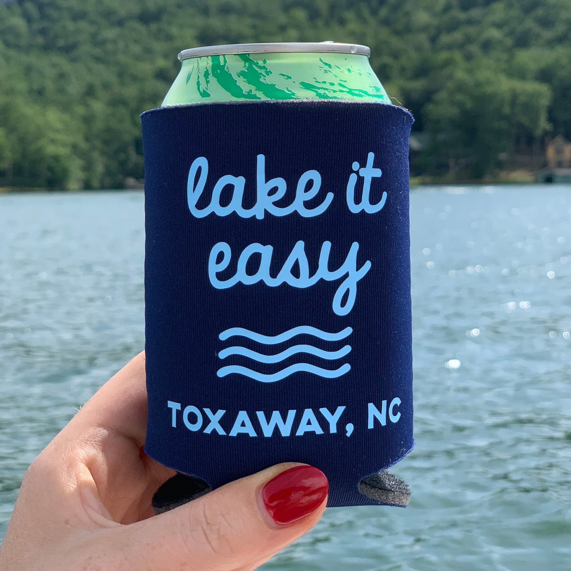 A person holds up a navy koozie which reads "lake it easy" and is customized with the city Toxaway, NC.
