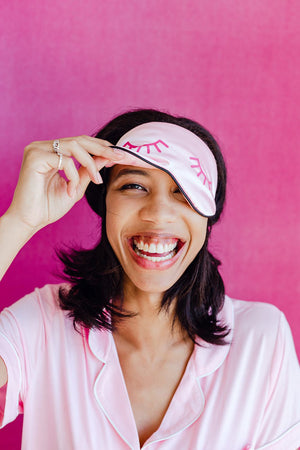 A woman wears a sleep mask with a bright pink eyelash design on it