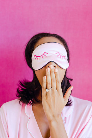 A woman wears a sleep mask with a bright pink eyelash design on it